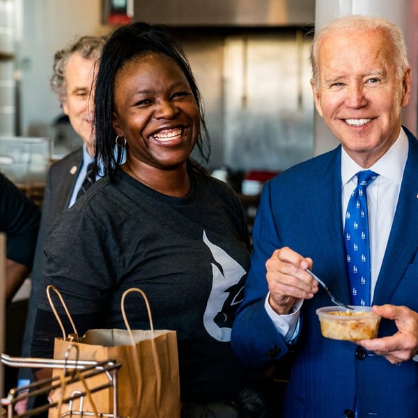 Joe Biden smiles, holding a food container, with two smiling restaurant staff members behind him.