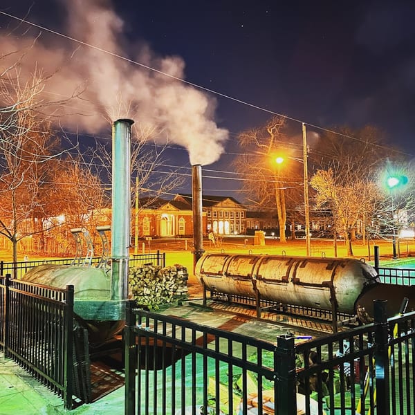 Two large barbecue smokers with smoke rising into the night sky, surrounded by a fence.