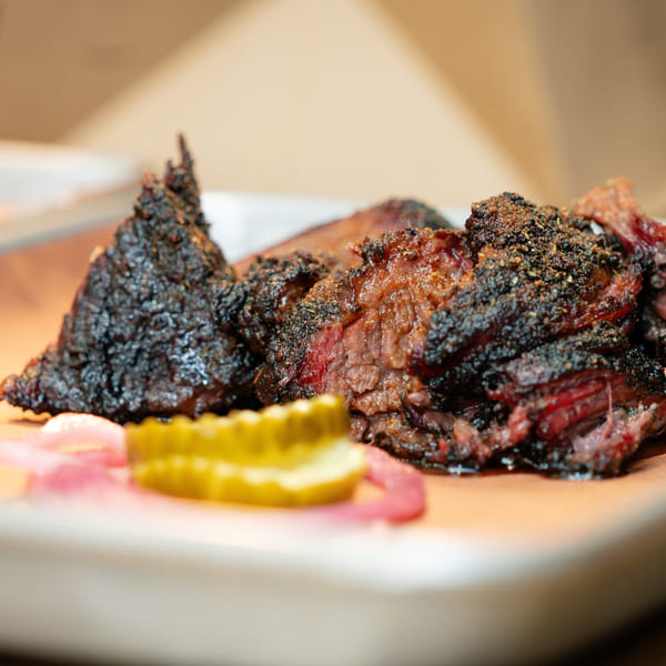Close-up of juicy, smoked brisket with a dark crust, served with pickles on a tray.